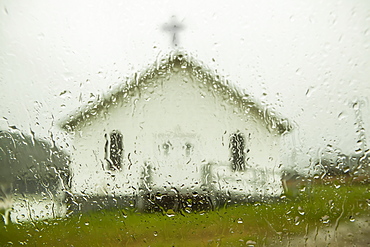 A white church building with cross viewed through a window wet with raindrops, Newfoundland, Canada