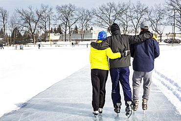 Two Males And One Female Skating Arm In Arm On Freshly Groomed Ice On Pond With Community Centre In The Background, Calgary, Alberta, Canada