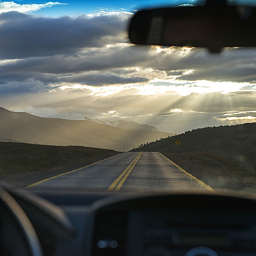 Rays Of Sunlight Shining Through The Clouds Onto The Road As Viewed Through The Windshield Of A Vehicle, Patagonia, Chile