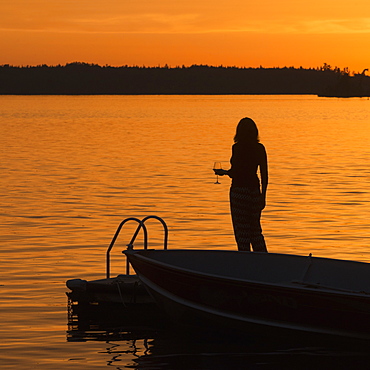 Silhouette Of A Woman Holding A Glass Of Wine While Standing On A Dock On A Golden Lake At Sunset, Lake Of The Woods, Ontario, Canada