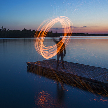 A Man Stands On The End Of A Dock At Sunrise Moving A Glowing Light In A Circular Form, Ontario, Canada