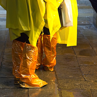 People Standing With Yellow Rain Ponchos And Footwear Covered In Orange Plastic Covers, Venice, Italy