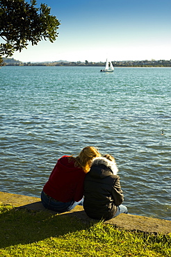 Two Girls Sitting On A Retaining Wall At The Water's Edge, Auckland, New Zealand