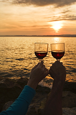 A couple holding wine glasses up at sunset over a lake, Bardolino veneto italy