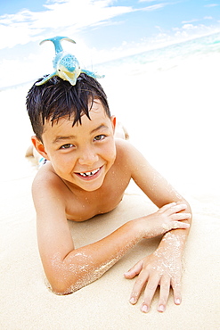 A Boy Laying On The Sand At The Water's Edge With A Toy Whale On His Head, Kailua, Oahu, Hawaii, United States Of America