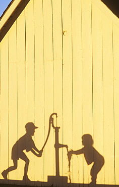 Agriculture - Silhouette on the side of a barn in late afternoon light of a farm boy operating a hand pump water well while his young sister washes her hands / Northwest Missouri, USA.