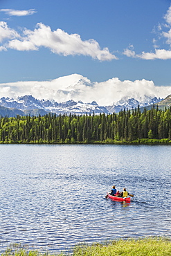 Two Women And Young Girl In A Red Canoe On Byers Lake With Green Forested Shoreline And Mount Mckinley Peaking Through The Clouds, Denali State Park, Alaska, United States Of America