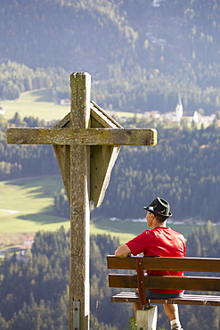 Male Hiker Sitting On Bench With Large Wooden Cross Overlooking Valley With Gren Meadows And Trees, Aschau, Austria