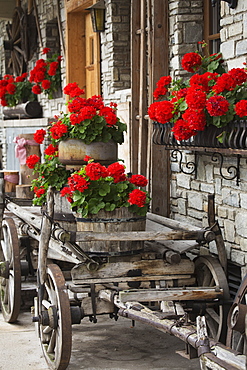 A Wooden Cart With Barrels Full Of Red Flowers Next To A Stone Building With Full Flower Boxes, Lanserbach, Austria