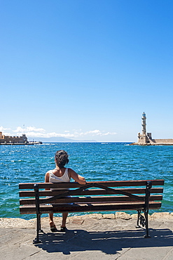 A Woman Sits On A Bench Overlooking The Harbour And Lighthouse, Chania, Crete, Greece