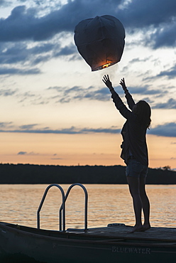 Silhouette Of A Girl Releasing A Lit Paper Lantern Into The Air At Sunset, Lake Of The Woods, Ontario, Canada