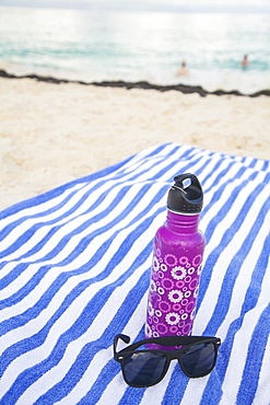 A Water Bottle And Sunglasses Sit On A Beach Towel On The Sand With View Of Swimmers In The Ocean, Oranjestad, Aruba