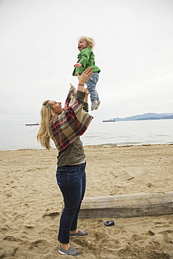 In A Winter Beach Scene, A Mother Joyfully Throws Her Little Girl In The Air, Vancouver, British Columbia, Canada