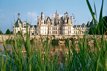 A grand chateau on a riverbank seen through grass, Chambord, France