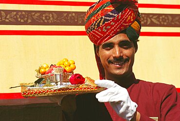 Portrait of a train steward holding winter cherries and a Rose flower on a serving tray, India