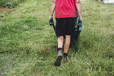 Rear view of farmer walking along field, pushing wheelbarrow.