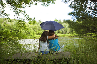 Two children, girls sitting together by a lake, under an umbrella, New York state, USA