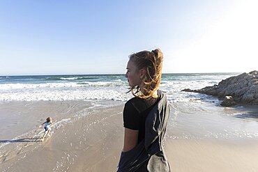 Teenage girl standing looking out over a beach, a boy running on the sand below, Grotto Beach, Western Cape, South Africa