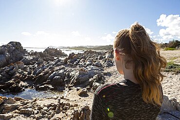 Teenage girl looking out over a beach and jagged rocks to the ocean, Pearly Beach, Gansbaai, Western Cape, South Africa