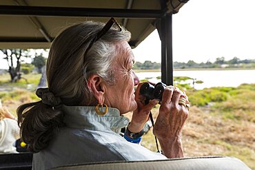 Senior woman using binoculars, sitting in a safari vehicle, looking out over marshes and waterway, Okavango Delta, Botswana