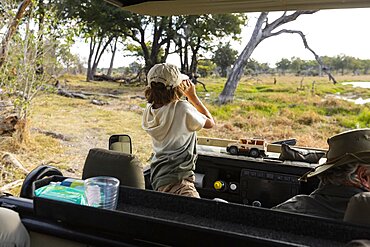 Young boy using binoculars standing up in a safari jeep, looking over the landscape, Okavango Delta, Botswana