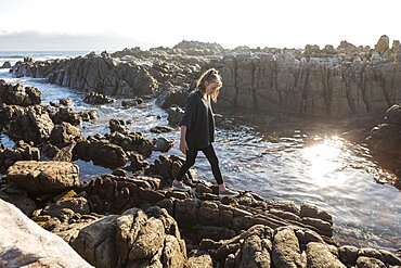 Teenage girl walking across jagged rocks, exploring rock pools by the ocean, De Kelders, Western Cape, South Africa