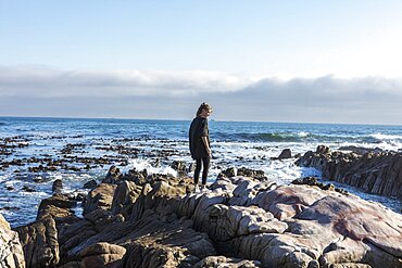 Teenage girl walking across jagged rocks, exploring rock pools by the ocean, De Kelders, Western Cape, South Africa