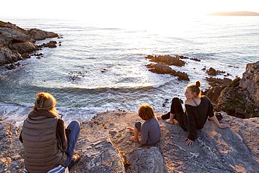 A family, mother and two children sitting watching the sunset over the ocean, Walker Bay Nature Reserve, South Africa