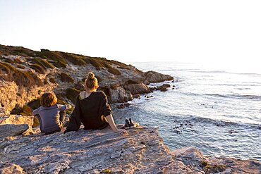 Teenage girl and young brother at sunset, sitting side by side looking over the ocean, Walker Bay Nature Reserve, South Africa