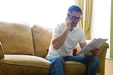 Man sitting on sofa making phone call holding paperwork.