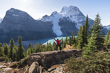 Man with arms raised, on a ridge above Berg lake and mountains.