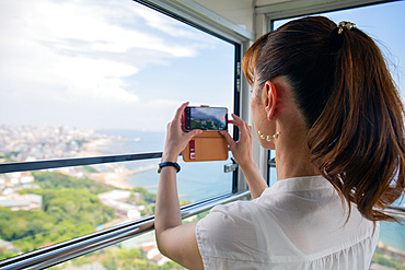 A mature Japanese woman using her mobile phone to take pictures from a cable car cabin of the city and landscape below.