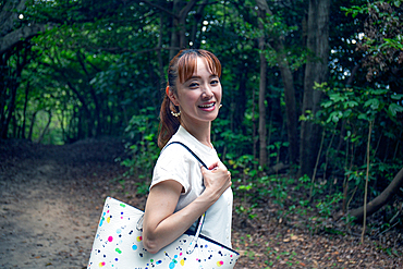 A mature Japanese woman outdoors in a park, head turned smiling at the camera.