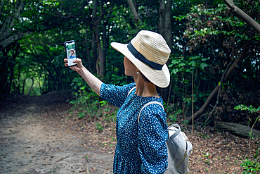 A mature Japanese woman in a straw hat and blue dress taking a selfie with a mobile phone.