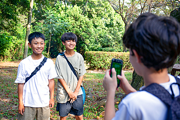 A boy with a mobile phone taking a picture of two 13 year old boys side by side, outdoors in a park in summer.