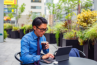 A young businessman in the city, on the move, a man seated at a cafe table outdoors, using a laptop, sipping a soft drink with a straw.