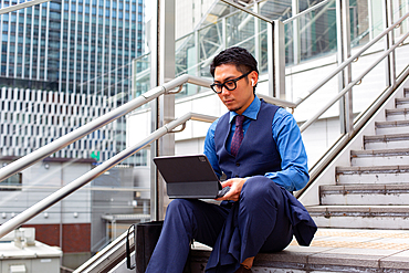 A young businessman in the city, on the move, seated on the stairs by a window, using a laptop.