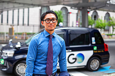 A young businessman in the city, on the move, a man with eyeglasses, shirt and tie, a taxi behind him.