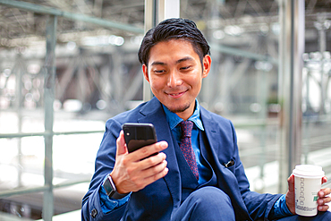 A young businessman in a blue suit in a city, looking at his mobile phone screen, texting or reading a message.