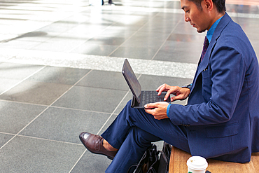 A young businessman in a blue suit on the move in a city downtown area, sitting on a bench using a digital tablet with a screen.