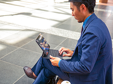 A young businessman in a blue suit on the move in a city downtown area, sitting on a bench using a digital tablet with a screen.