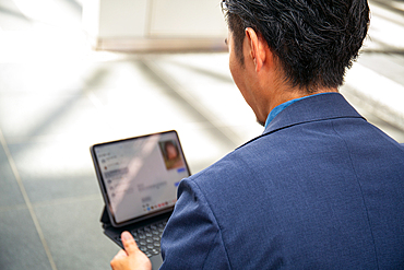 A young businessman in a blue suit on the move in a city downtown area, sitting on a bench using a digital tablet with a screen.