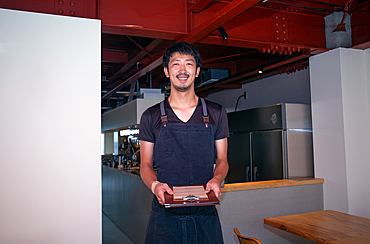 A man in a restaurant wearing an apron, holding a clipboard and menu, smiling in greeting.