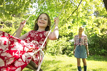 Summer. A girl in a sundress on a swing under a leafy tree. A woman standing behind her.