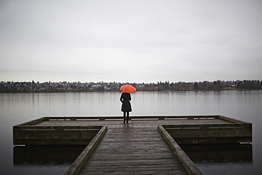 A woman wearing all black and holding an orange umbrella stands on the edge of a dock on a grey and cloudy day in Seattle, WA, Seattle, Washington, USA