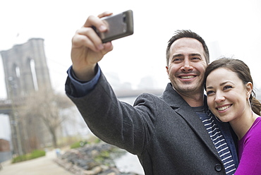 New York city. The Brooklyn Bridge crossing over the East River. A couple taking a picture with a phone, a selfy of themselves, New York city, USA