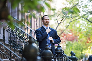 A man in a business suit adjusting his tie. At the bottom of the steps of a townhouse in a terrace.