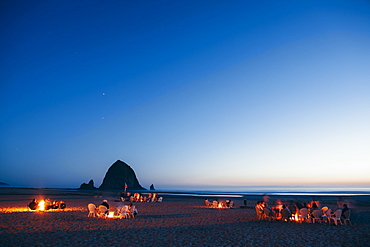 Crowds of people sitting by glowing campfires on Cannon Beach at dusk, Haystack Rock in the background, Tillamook County, Oregon, USA