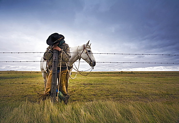 A cowboy standing leaning on a fence post on the range, A grey horse behind him, Saskatchewan, Canada