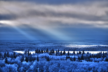 View over the landscape of pine forests, and snowfields and shafts of sunlight reflecting on the surface of a lake, Canada